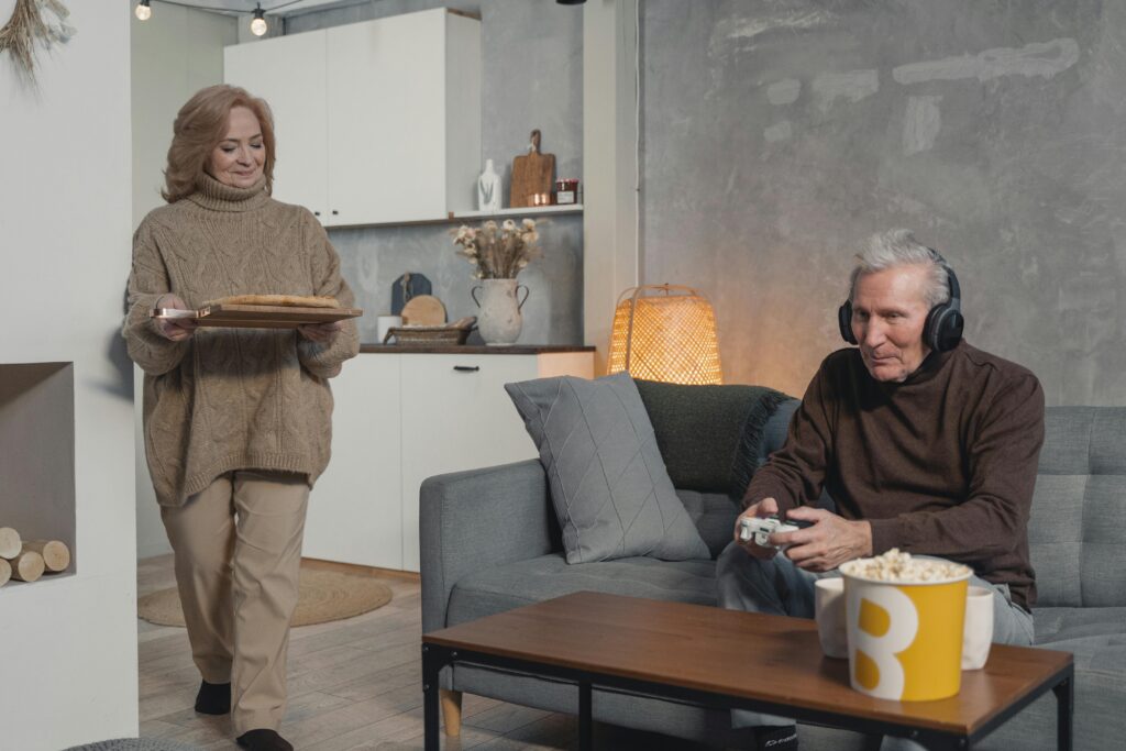 Senior couple having fun at home, with man gaming and woman serving food. Cozy indoor setting.